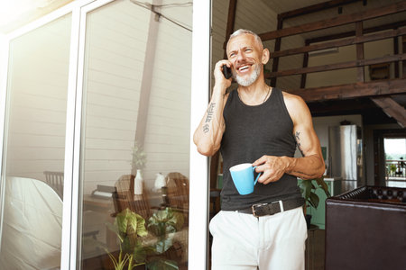 Good news. Happy middle aged caucasian man talking by mobile phone while drinking coffee near balcony of his modern flatの写真素材