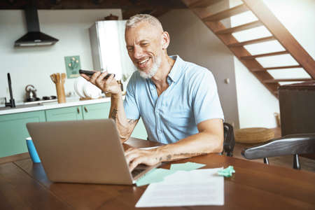 Good talk. Happy middle aged caucasian man talking by phone while while sitting at table and working online on laptopの写真素材