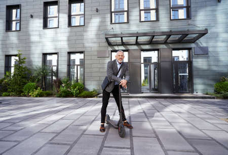 Moving forward. Full length shot of stylish mature businessman looking at camera while standing with electric scooter near modern office building outdoorsの写真素材