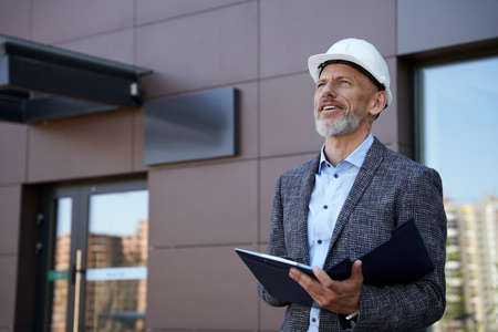 Monitoring the process. Confident middle aged architect, engineer manager wearing helmet looking away while standing outdoors and holding a blueprintの写真素材