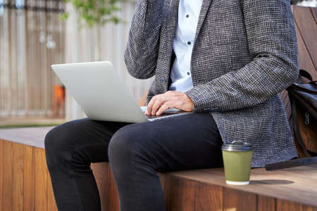 Cropped shot of busy elegant mature man talking on the phone, working on his laptop while sitting on the bench outdoorsの写真素材
