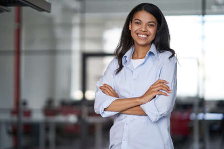 Portrait of a young happy attractive business woman or female office worker keeping arms crossed, looking at camera and smiling while working in the modern officeの写真素材