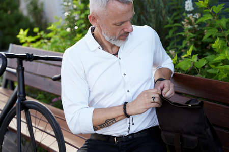 Modern middle aged businessman opens up his backpack, sitting on the bench outdoors with a bicycle next to himの写真素材