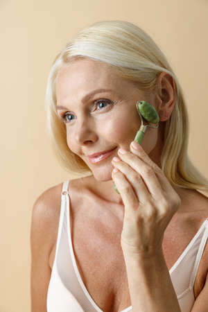 Close up portrait of mature blonde woman with glowing skin smiling aside while using jade facial roller for skin care and beauty treatment, posing isolated over beige backgroundの写真素材