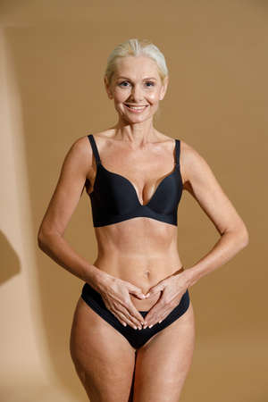 Studio shot of smiling mature blonde woman in black underwear holding hands on her belly, forming a heart symbol while standing isolated over beige backgroundの写真素材