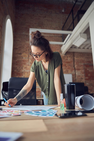 Creative woman, interior designer or architect wearing glasses making notes with a pen while working on a blueprint for new project, standing in her officeの写真素材