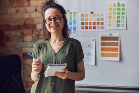 Attractive woman, interior designer or architect in casual wear with messy hairdo holding pen and notebook, smiling at camera, working on new creative project, standing near board with color samplesの写真素材
