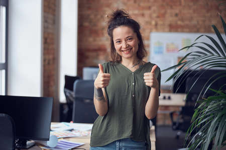 Portrait of cheerful woman, interior designer or architect smiling at camera, showing thums up while standing in modern green office on a daytimeの写真素材