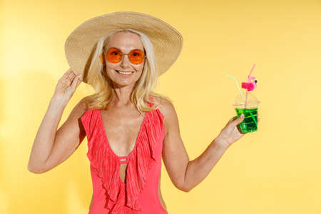 Cheerful blonde woman in sunglasses, red swimsuit and straw hat smiling at camera, adjusting her hat and holding cocktail drink while posing isolated on yellow backgroundの写真素材