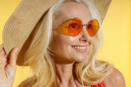 Portrait of happy blonde woman in sunglasses and straw hat smiling aside, adjusting her hat while posing isolated on yellow backgroundの写真素材