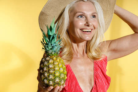 Portrait of dreaming blonde woman in red swimsuit and straw hat smiling away, holding fresh...
