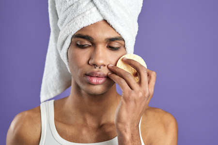 Close up portrait of handsome young man cleaning skin with cotton pads on isolated purple backgroundの写真素材