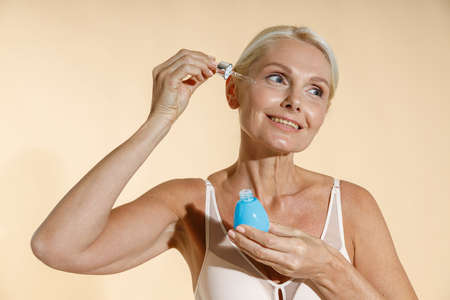 Studio shot of beautiful smiling mature woman looking aside while applying serum with dropper on her face, posing isolated over beige backgroundの写真素材