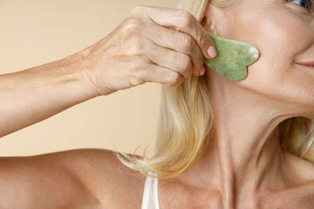 Close up shot of mature blonde woman with perfect glowing skin using jade Gua Sha massage tool for skin care and beauty treatment isolated over beige backgroundの写真素材