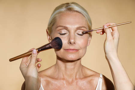 Close up of hands of skillful artist doing natural makeup for mature blonde woman isolated over beige background. The woman closed her eyes with serenityの写真素材