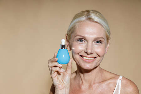 Beauty portrait of smiling mature blonde woman with bottle of serum posing isolated over beige backgroundの写真素材