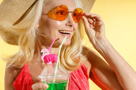 Close up portrait of cheerful blonde woman in sunglasses, red swimsuit and straw hat smiling aside,...
