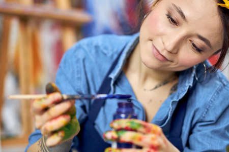 Portrait of young woman, female painter in apron holding blue paint jar and a paintbrush, sitting on the floor at home workshopの写真素材