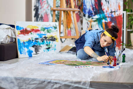 Talented female painter in apron choosing paint color, holding paintbrush while working on painting,...