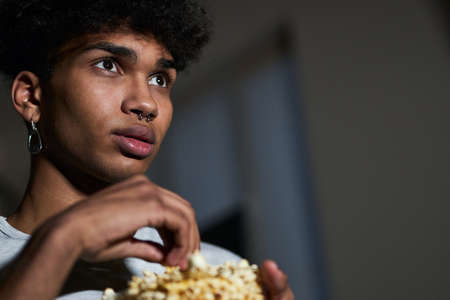 Close up portrait of young guy taking some popcorn from a bowl while watching movie at homeの写真素材