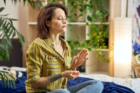 Portrait of calm young woman in casual wear sitting in Lotus pose, doing some meditation at homeの写真素材