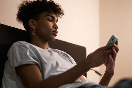 Social networks. Portrait of young guy with piercing using smartphone in his bed at night before going to sleepの写真素材