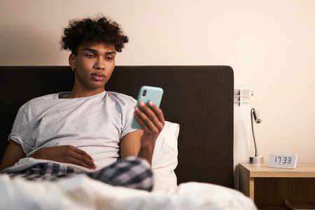 Social media addiction. Young guy with piercing reading news, using smartphone in his bed at night before going to sleepの写真素材