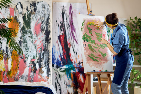 Back view of talented female artist working on a modern abstract oil painting. She is applying paint on canvas with fingers, standing in home studio workshopの写真素材