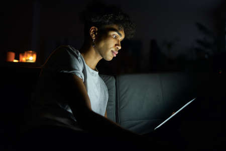 Side view of focused young guy reading e book or watching video using tablet pc at night, sitting in dark room at homeの写真素材