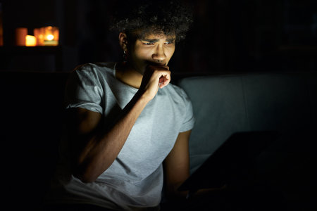 Portrait of young guy looking concentrated while reading e book using tablet pc at night, sitting in dark room at homeの写真素材