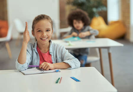 Active student. Cute little school girl smiling at camera raised her hand while sitting at the desk in classroomの写真素材