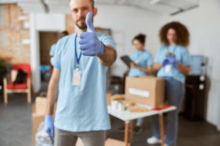 Male volunteer in blue uniform and protective gloves showing thumbs up at camera. Team sorting, packing items in cardboard boxesの写真素材