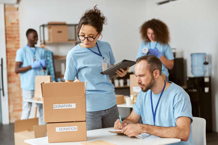 Man and woman working together on donation project indoors. Team of volunteers in blue uniform making notes, calculating, sorting and packing items in cardboard boxesの写真素材
