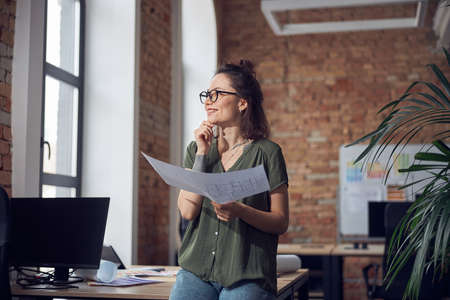 Smiling creative woman, interior designer or architect wearing glasses looking away, working on a blueprint for new project, standing in her officeの写真素材