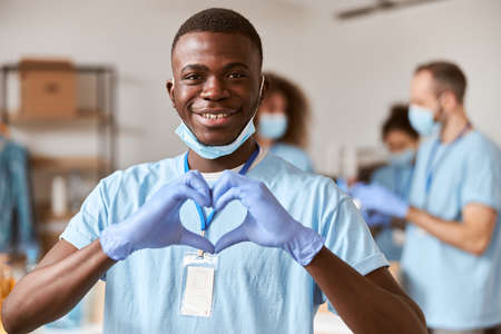 Closeup portrait of african american young male volunteer in blue uniform, protective mask and gloves smiling at camera and showing heart love sign. Team sorting, packing food stuff in the backgroundの写真素材