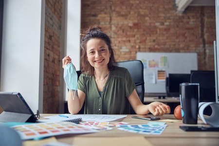 Smiling woman, interior designer holding protective mask and looking at camera, working with color samples for new project, sitting at the table in her officeの写真素材