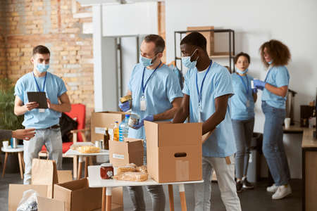 Diverse volunteers in blue uniform, protective masks and gloves separating donations stuffs together, sorting items in cardboard boxes while standing indoorsの写真素材