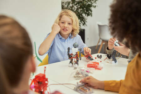Lively little caucasian boy looking emotional while sitting at the table and examining details of a robot during STEM classの写真素材