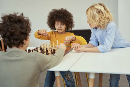 Awesome little boy with afro hair looking concentrated while playing chess in the room with other childrenの写真素材