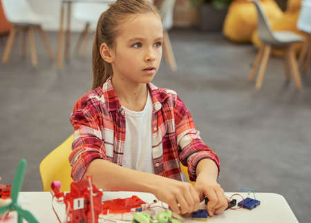 Beautiful little caucasian girl looking away while putting an electronic toy kit together, sitting at the table during STEM classの写真素材