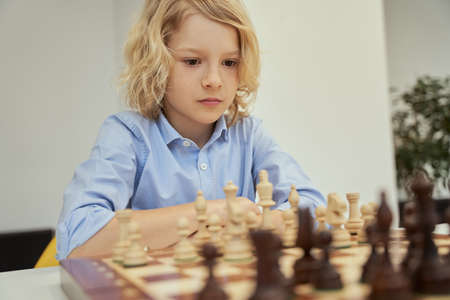 Concentration. Portrait of focused caucasian boy in blue shirt sitting in the classroom and thinking while playing chess on the chessboardの写真素材