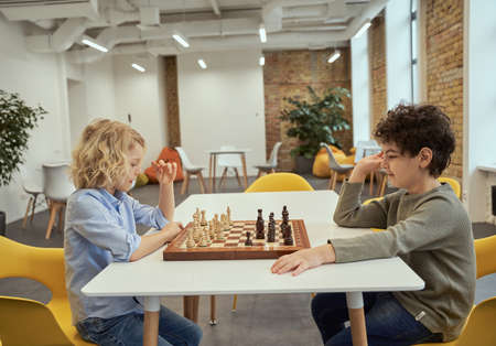 Mind sport. Side view of two smart little boys playing chess while sitting at the table in schoolの写真素材