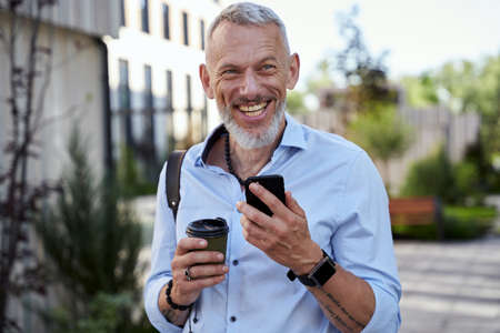 Happy modern middle aged businessman smiling while using his smartphone, walking outdoors with a cup of coffeeの写真素材