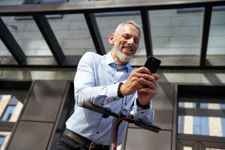 Generating ideas. Cheerful middle aged businessman smiling while using his smartphone, standing with electric scooter outdoors near modern buildingの写真素材