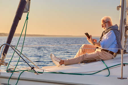 Side view of a retired man sitting on the side of his sailboat or yacht floating in the calm blue sea at sunset, using digital tablet and relaxingの写真素材