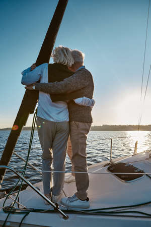 Rear view of senior couple hugging and looking at the horizon, enjoying amazing sunset while standing on the side of sail boat or yacht deck floating in seaの写真素材