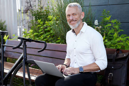 Building a Business. Successful modern middle aged businessman in stylish suit smiling at camera while working on his laptop, sitting on the bench outdoors with a bicycle next to himの写真素材