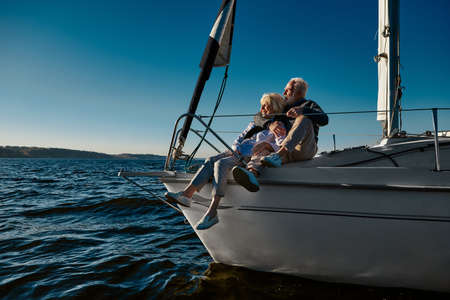 Happy retired family couple relaxing on a sail boat or yacht deck floating in a calm blue sea, hugging and enjoying amazing viewの写真素材