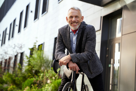 Confident, stylish mature businessman smiling at camera while standing with his bicycle outdoors on a daytimeの写真素材