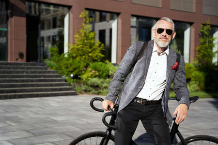 Commuter. Confident, stylish middle aged business man in sunglasses standing with his bicycle near his workplace and looking at cameraの写真素材